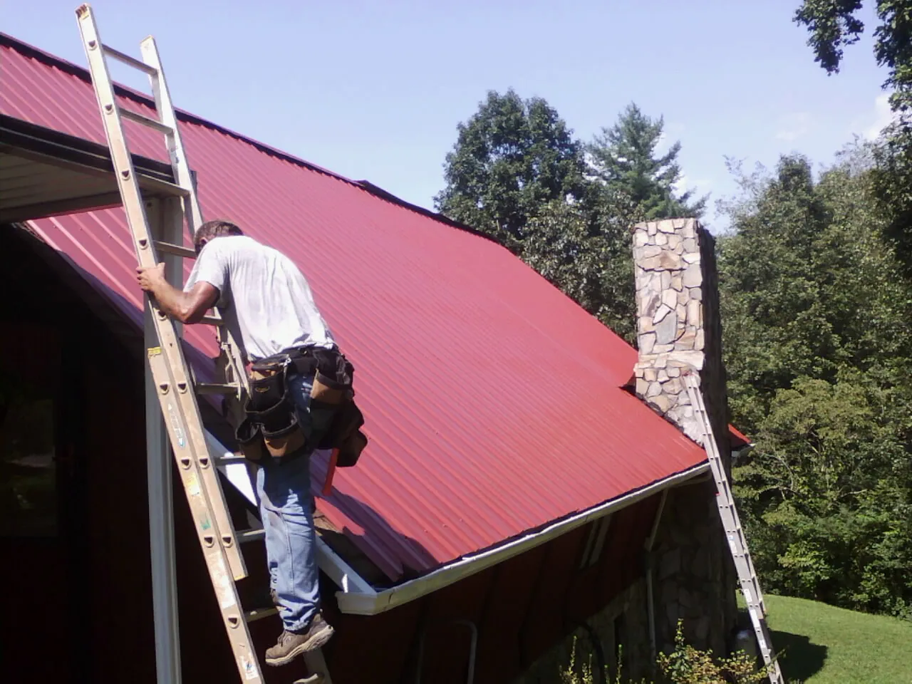 Roofer on a ladder beside a red metal roof and stone chimney — Soffit & Fascia Repair by Roofing Brothers in Blodgett Mills